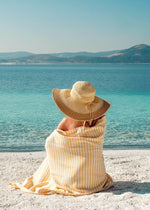 girl wrapped in Turkish towel with stripes and tassels, sitting on beach in straw hat and Bezzazan Harmonious beach towel