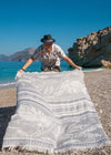 Flourishing unisex eco-friendly turkish towel by Bezzazan, with white fringe and palms, held by smiling male model at beach